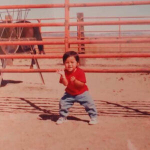 An old photo of a young child outside of a pen for horses or cows. They have dark hair, and wear a red shirt and baggy pants. The pen is empty, except for an obscured, inanimate object. The landscape is dry and dusty.