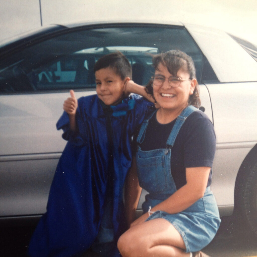 An old photo of a young child and an adult smiling for the camera in front of a silver car. The child has a graduation robe on, and leans familiarly on the adult. The adult kneels down to the child's height. They wear denim overalls over a black t-shirt. 