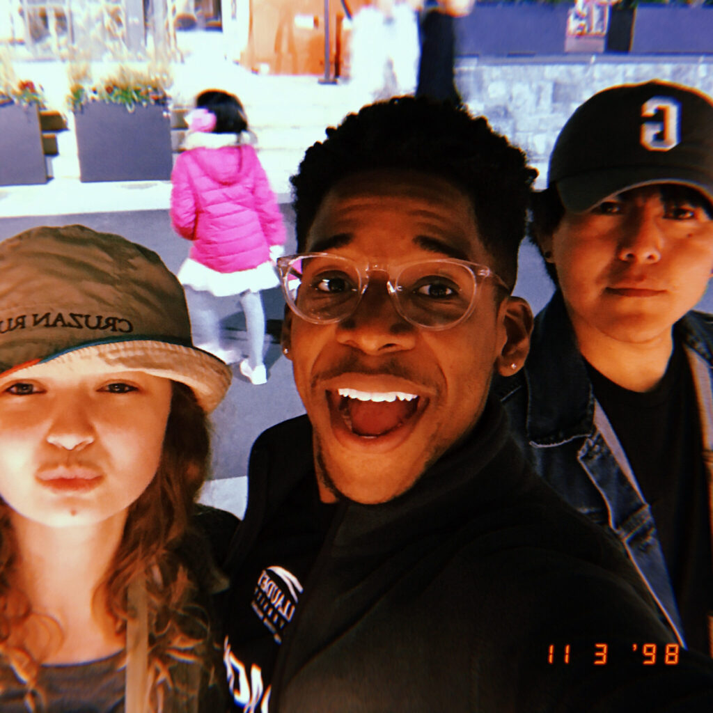 A photo of three friends taking a selfie. The person the left, wearing a bucket hat, is pursing their lips. The person in the middle is holding the camera and smiling brightly. The person on the left, wearing a cap, has a neutral look on their face.