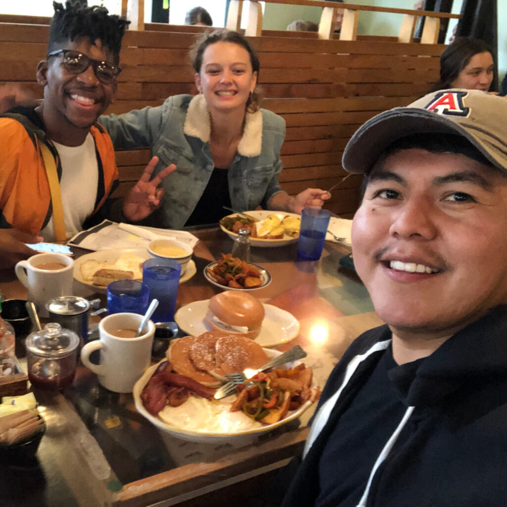 A photo of three friends smiling for a selfie at a restaurant table. The person holding the camera, on the right, is wearing a University of Arizona cap. The person in the middle has a denim jacket with soft lining. The person on the left has glasses, tall styled hair, and makes a peace sign.