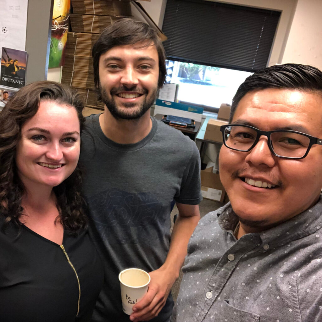 A photo of three friends taking a selfie. The person the left has long, dark hair and a black shirt. The person in the middle has short dark hair and a short beard, and holds a paper cup that reads "Be Fab!". The person on the left, holding the camera, has styled black hair and a light goatee.