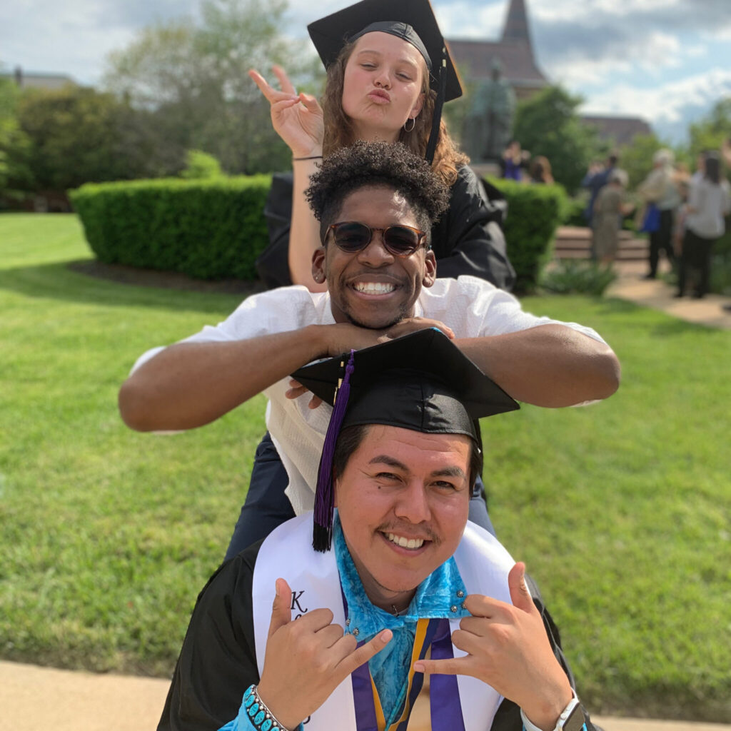 A photo of three friends smiling posing for a photo outside of a graduation ceremony. They are playfully stacked vertically, one head over another. The people on top and bottom have graduation caps. The person on top purses their lips. The person in the middle has sunglasses. The person on bottom makes a shaka hand gesture.