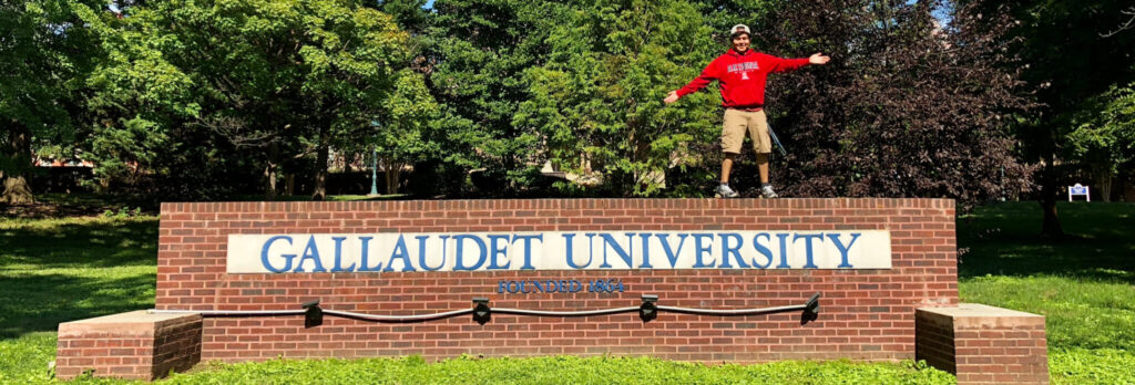A photo of a student in red, center right, standing with arms outstretched on a "Gallaudet University" sign on the school's campus. The name is enclosed within brick. In the background are trees, with a building tower peaking above the treeline in the top center left.