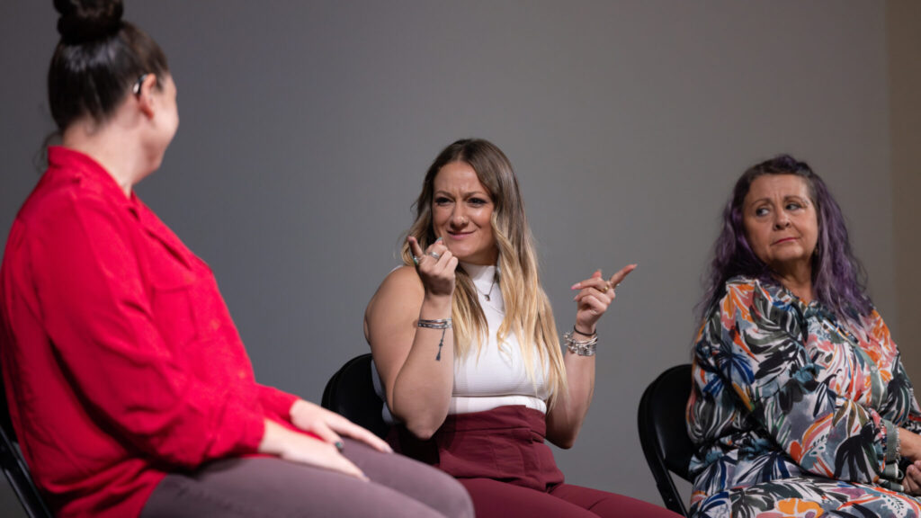 A photo of a panelist signing onstage, looking at a moderator out of focus in the left foreground The panelist has brown and blonde hair, a white sleeveless top, and brown pants. on the right is another panelist, watching.