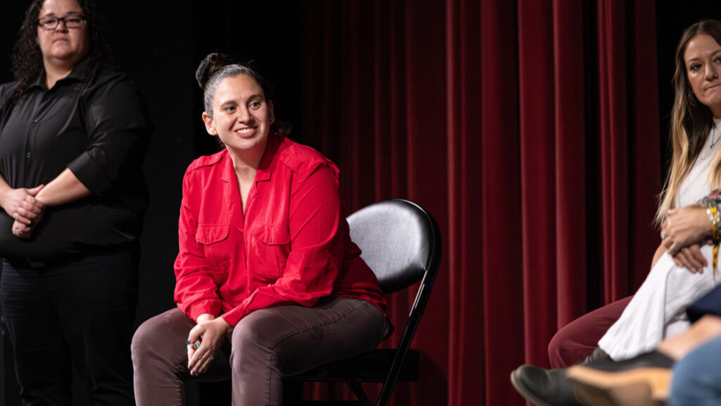 A photo of a seated person in red onstage, looking toward panelists who are mostly offscreen to the right, except for one on the far right of the image. The person in red has a bright red formal shirt, and dark hair in a bun behind their head. They are leaning forward attentively and smiling. An interpreter is visible behind them, watching.