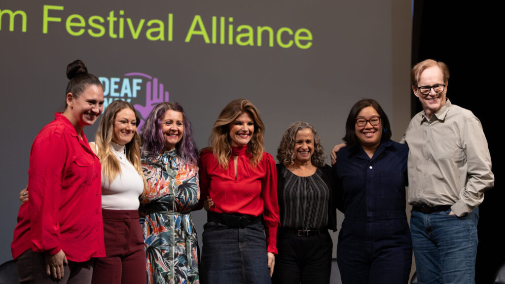 A photo of seven people onstage, looking down at an unseen camera to the bottom right. They are wearing various outfits of playful formal attire. Behind them, a screen has obscured words that include "Festival Alliance" and the Deaf Way logo.