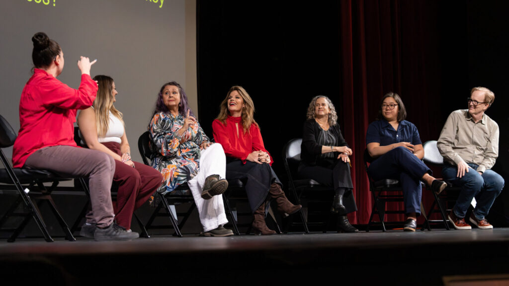 A photo of seven people onstage, sitting in folding chairs. On the right, a moderator in red addresses the panelist third from right. The other panelists look on.