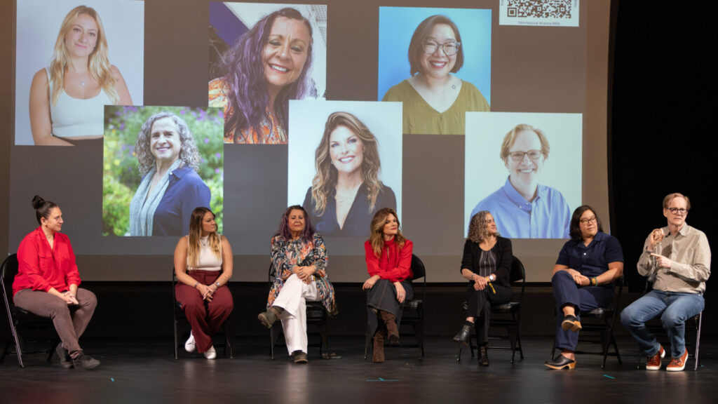 A photo of seven people onstage, sitting in folding chairs. The six on the left are looking towards the person on the far right, signing to a crowd offscreen. Behind them is a projected screen with photos of the six panelists on the right.