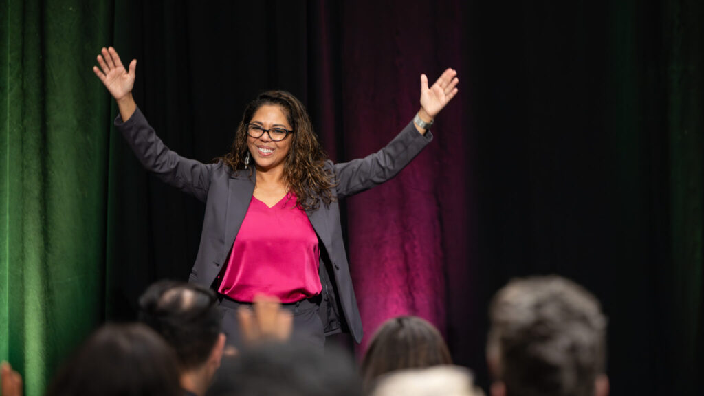 A photo of a performer looking to a crowd behind the camera, with some heads out of focus in the foreground. They are raising their arms in welcome. They have dark wavy hair and glasses. They wear gray pantsuit and a purple top.