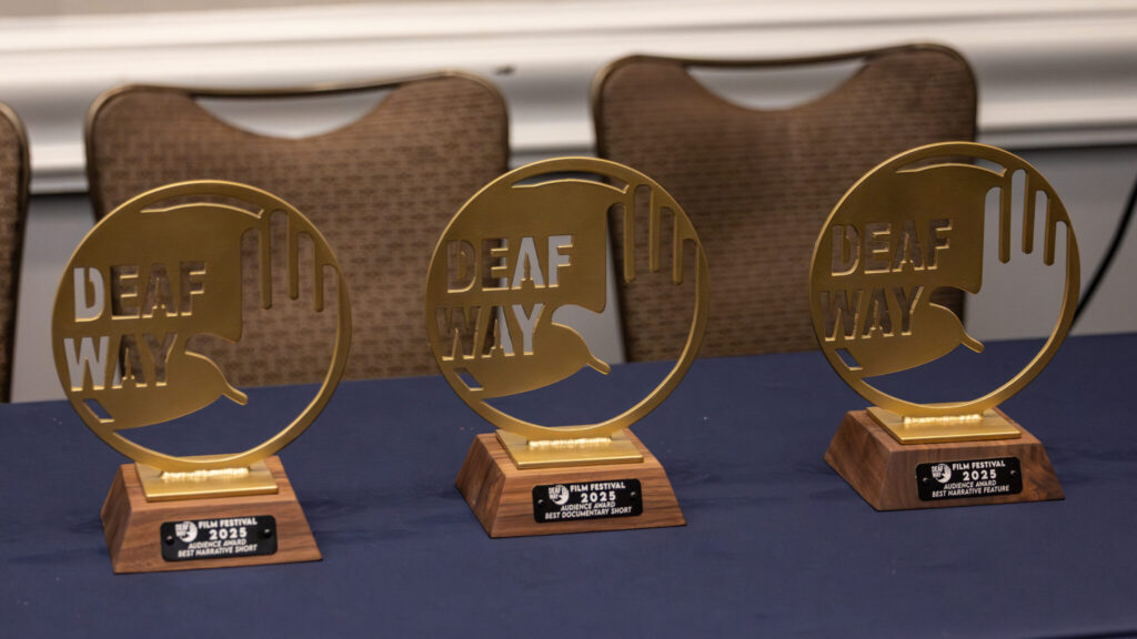 A photo of three identical circular golden awards on a table with a blue tablecloth. The awards have cutout of a logo that says "Deaf Way".