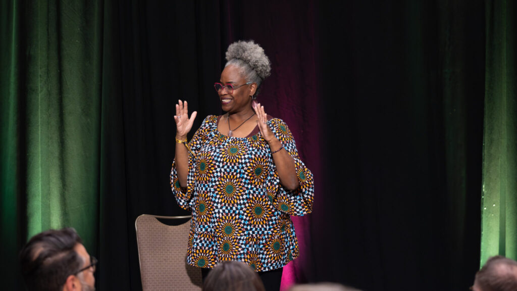 A photo of a person handwaving on a stage with a black background. They have curly gray hair in a big bun behind their head, glasses, and a patterned shirt with sunflowers.