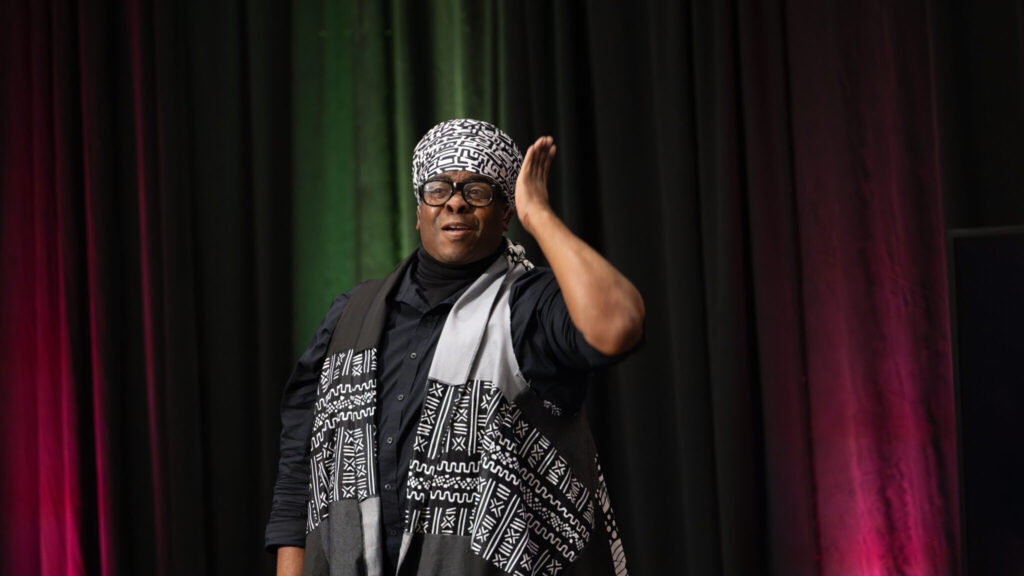 A photo of a performer looking to an unseen crowd behind the camera. They have a loving look and their left arm is up in performance. They wear a patterned head wrap and light vest in black and white over a black shirt.