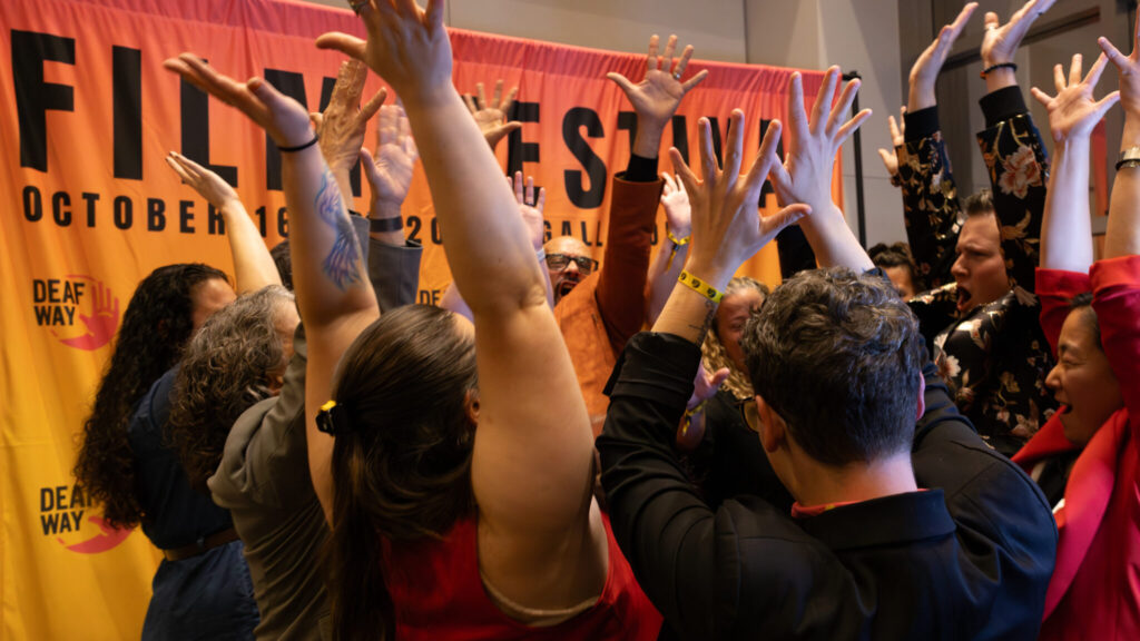 A photo of several people in a huddle raising their arms and cheering. Behind them is an orange screen that says "Film Festival / October 16-19, Gallaudet University".