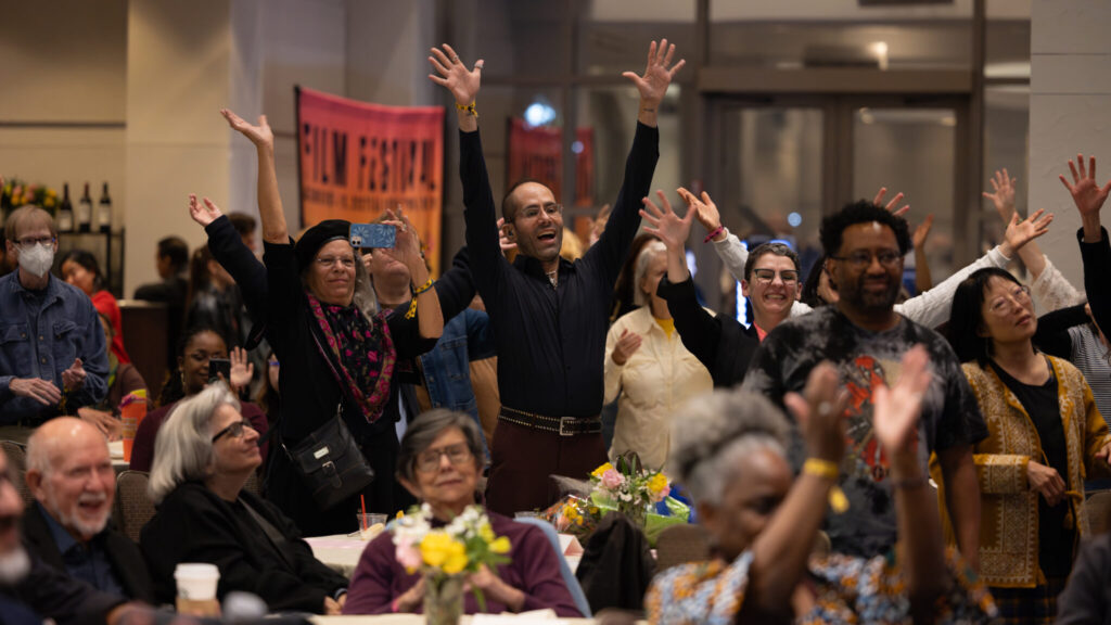 A photo of a crowd in a ballroom raising their arms and handwaving an unseen stage to the right behind the camera. In the foreground, out of focus, most are seated. In the background, many are standing. Prominent in the center is a taller individual with their arms outstretched.