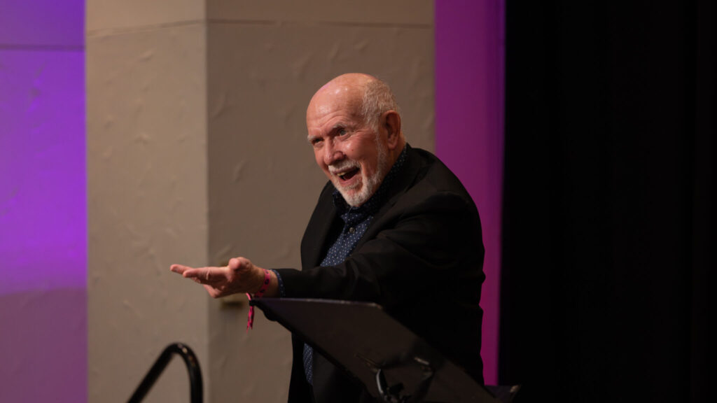 A photo of a performer looking to an unseen crowd to the left. They have a playful look and reach out with their left arm. They are bald, with white hair around the sides and a short beard. They wear a dark jacket and shirt.