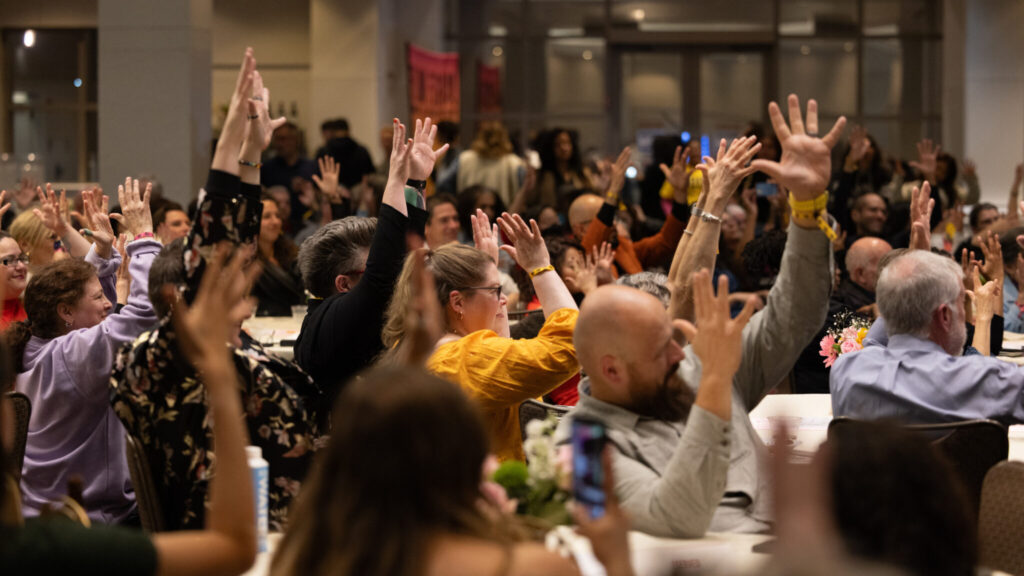 A photo of a seated crowd in a ballroom raising their arms and handwaving an unseen stage to the right.