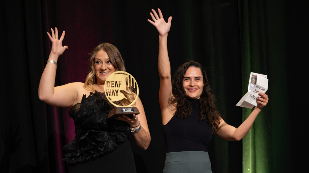 A photo of two award presenters handwaving on a stage. The person on the left is holding a circular golden award that says "Deaf Way". They have dark blonde hair and wear a feathery black dress. The person on the right has dark wavy hair, with a black top and grey bottoms.