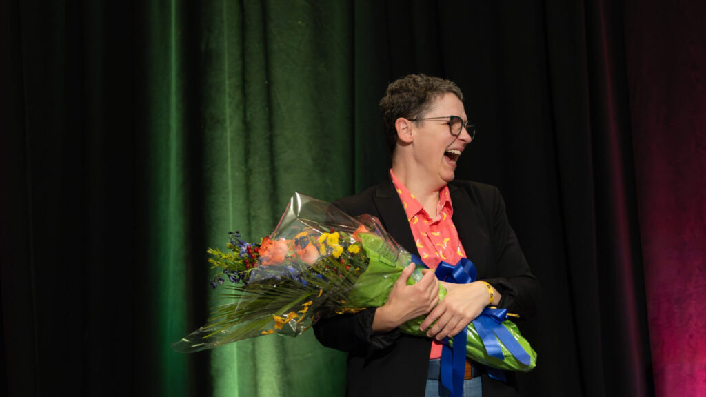 A photo of a person onstage holding a colorful bouquet and smiling brightly to somebody offscreen to the right. They have short hair and glasses. They wear a dark jacket and bright shirt.