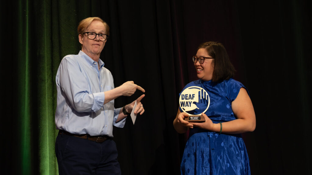A photo of two award presenters on a stage. The person on the right is holding a circular golden award that says "Deaf Way". They wear a striking blue dress and smile at the person on the left. The person on the left is tall, with a blue shirt and black pants. They are signing something towards the award.