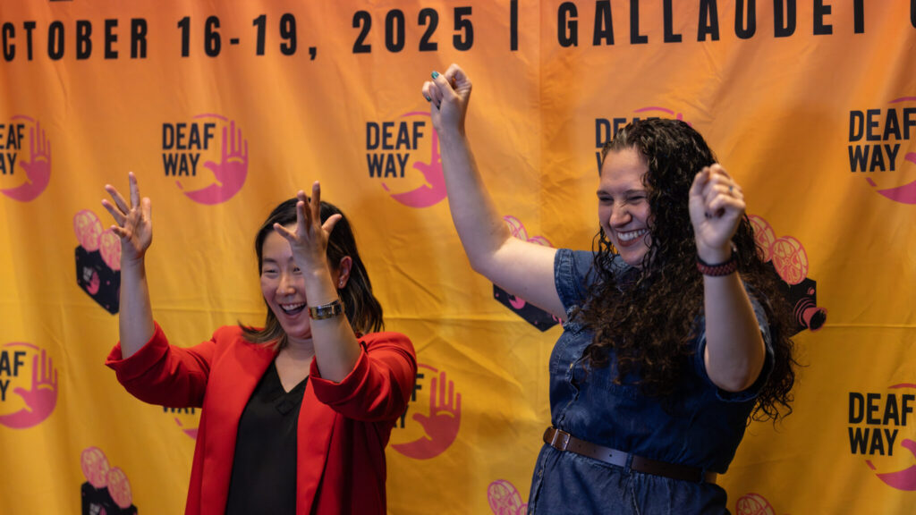 A photo of two smiling and cheering for a camera offscreen to the left. Behind them is an orange screen that says "October 16-19, Gallaudet" and has Deaf Way logos. The person on the left has a dark hair and a red jacket. The person on the right has long, dark wavy hair and a blue dress.
