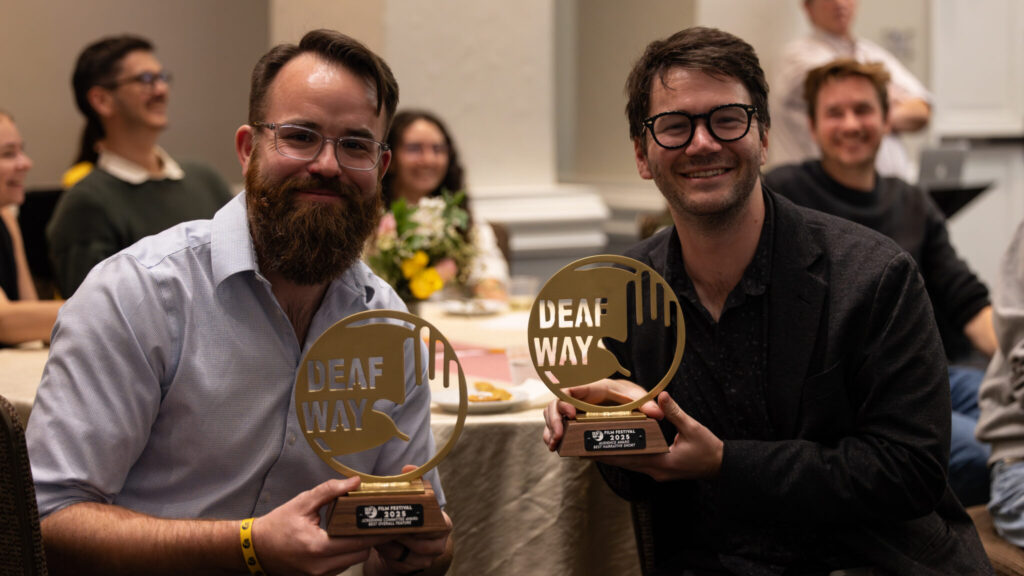 A photo of two seated people smiling for the camera and holding up circular awards that say "Deaf Way". The person on the left has brown hair and a thick beard, with glasses and a blue button-up shirt. The person on the right has tousled brown hair, with glasses and a dark jacket. More people are in the background around a table.