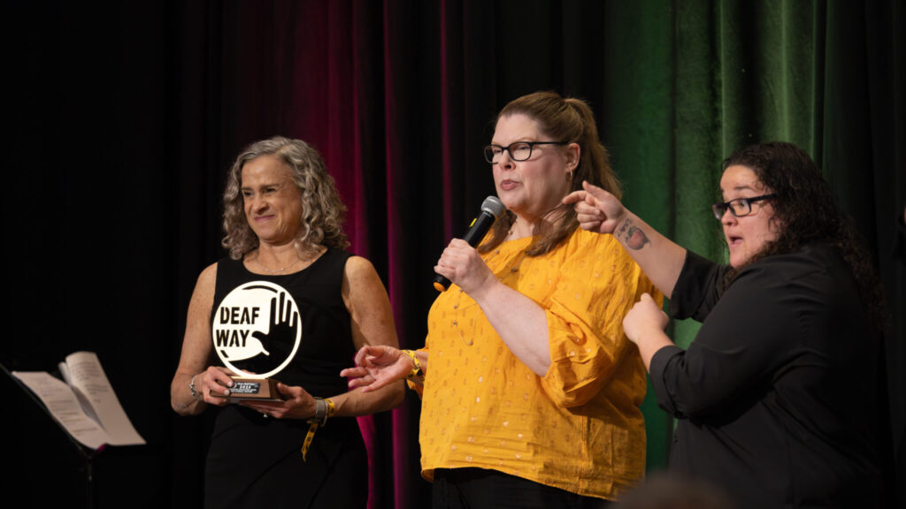 A photo of two award presenters on a stage with an interpreter. The person on the left is holding a circular golden award that says "Deaf Way". They wear a black dress and look at an unseen audience to the left. The person in the middle holds a microphone. They have glasses and a yellow top. The interpreter on the right is signing.