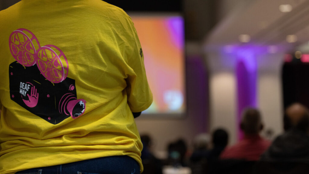 A photo from behind of a person's back, focusing on their yellow t-shirt. The shirt has a black and pink camera illustration with the Deaf Way logo.