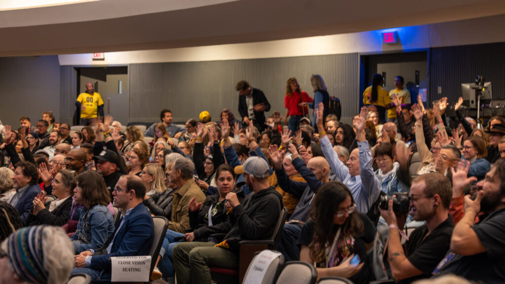 A photo of a large crowd in a theater. Many are cheering a stage offscreen to the left.