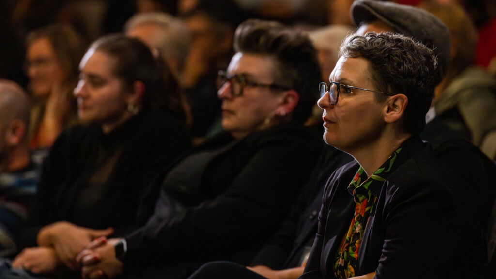 A photo of a seated audience. The person in focus is on the right, looking attentively to the unseen stage to the left, along with the out-of-focus observers behind.