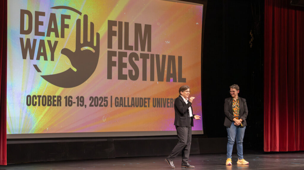A photo of two people onstage in front of a projected screen that has the Deaf Way logo and says "Film Festival / October 16-19, 2025 / Gallaudet University". The person on the left is signing to the unseen crowd to the right. The person on the right is smiling at the person on the left.