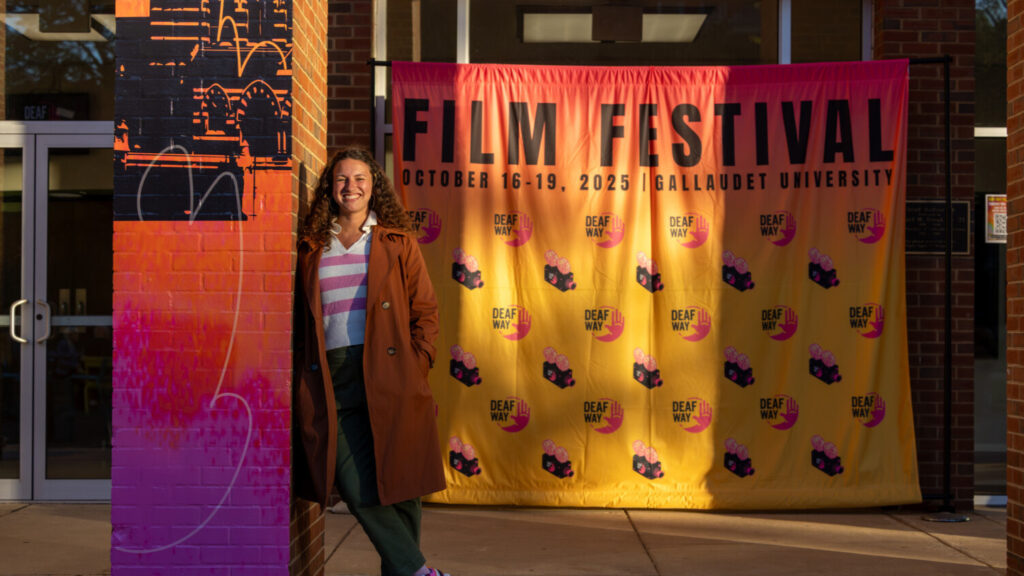 A photo of a person under dusky sunlight, standing in front of a building and leaning on a column. The column is decorated in colorful orange and purple. A backdrop in the background is also orange, and says "Film Festival / October 16-19 / Gallaudet University" with the Deaf Way logo. The person is smiling casually. They wear a long coat and striped white-and-pink shirt.