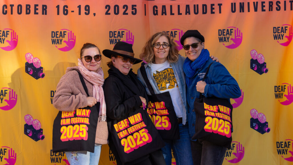 A photo of four people posing in front of an orange backdrop with the Deaf Way logo. They are all holding bags that say "Deaf Way Film Festival 2025".