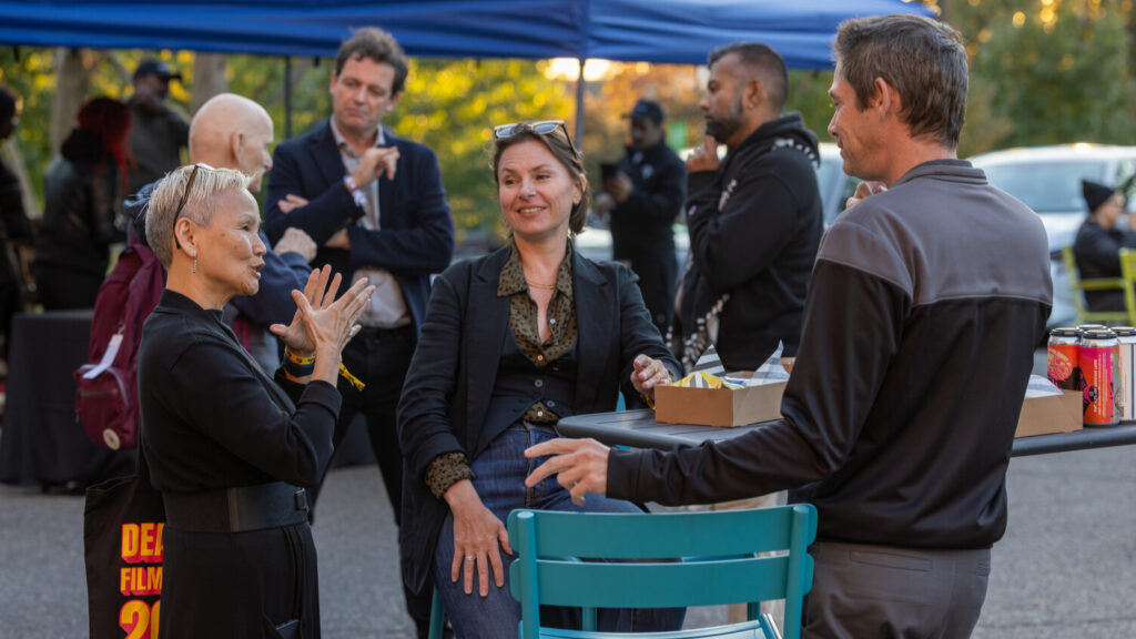 A photo of three people chatting in ASL around an outdoor cocktail table. The light indicates a shady area on a sunny day. The person on the left is signing, and the other two are looking. More people are out of focus in the background.