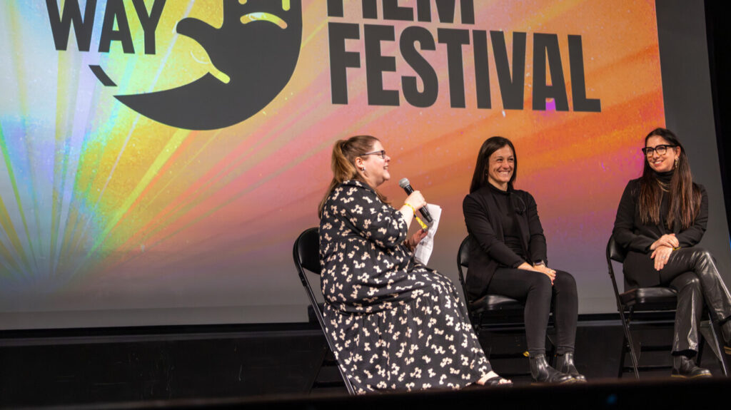 A photo of three seated people onstage. The person on the left is holding a microphone and speaking. They wear a long patterned dress. The person in the middle is sitting and smiling toward the unseen crowd. The person person on the right is looking at the speaking person and smiling. In the background is a projected screen with an obscured Deaf Way Film Festival logo.