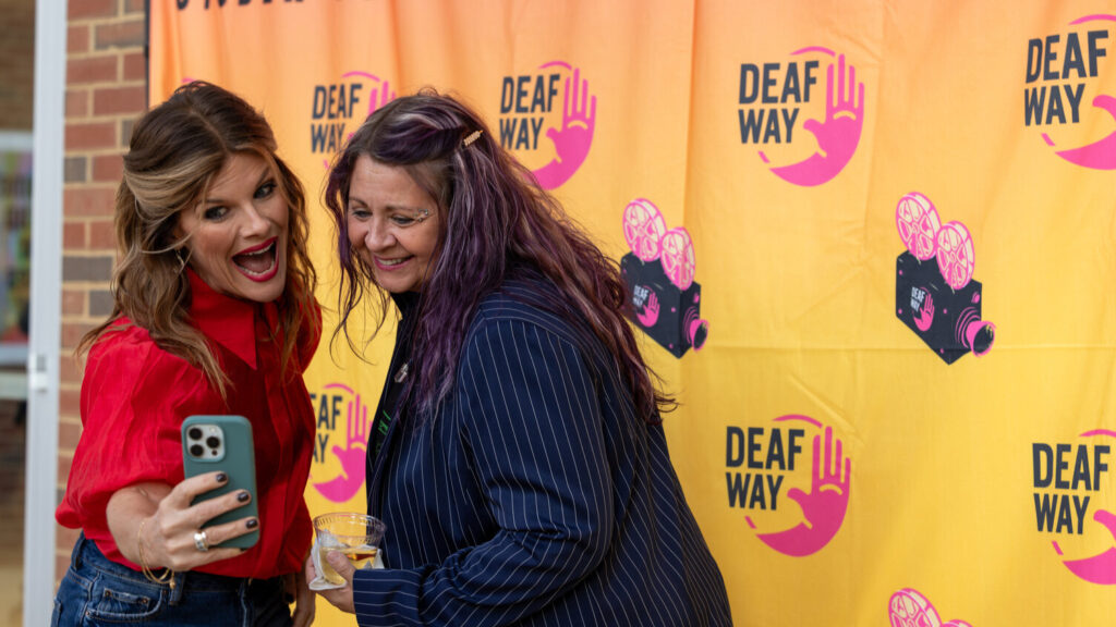 A photo of two people taking a selfie in front of an orange backdrop with the Deaf Way logo. The person on the left is holding the phone and making a playful face of cheer. The person on the right is smiling warmly toward the phone.