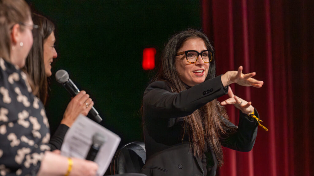 A photo of a person, center right, sitting onstage and gesturing to an unseen crowd to the right. They have long dark hair. They wear glasses and a dark coat. Their expression is engaging. On the left, obscured and out of focus, are two people. One is an interpreter holding up a microphone and watching the person on the right.