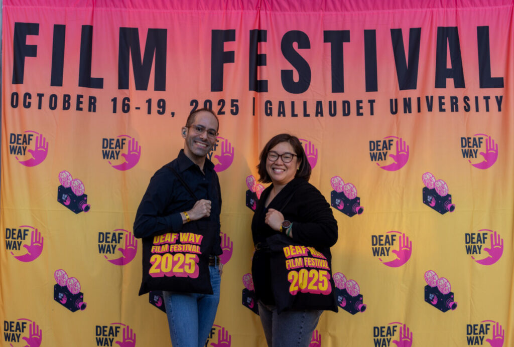 A photo of two people posing in front of an orange backdrop that says "Film Festival / October 16-19 / Gallaudet University" with the Deaf Way logo. They are holding black bags that say "Deaf Way Film Festival 2025".