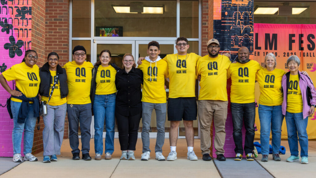A photo of eleven people in a single row posing for the camera with their arms around each other. All but one wear a yellow t-shirt with black text: "QQ / Ask me". The person without a t-shirt is wearing a black jumpsuit.