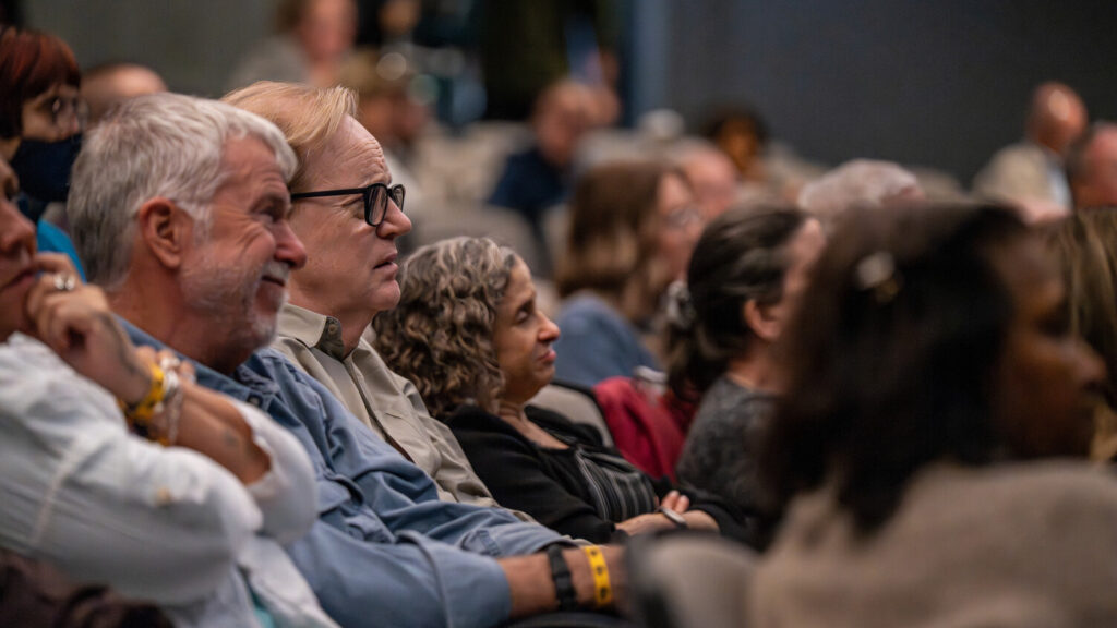 A photo of a seated audience in a theater. In focus are three people in the middle watching the unseen stage to the right thoughtfully.
