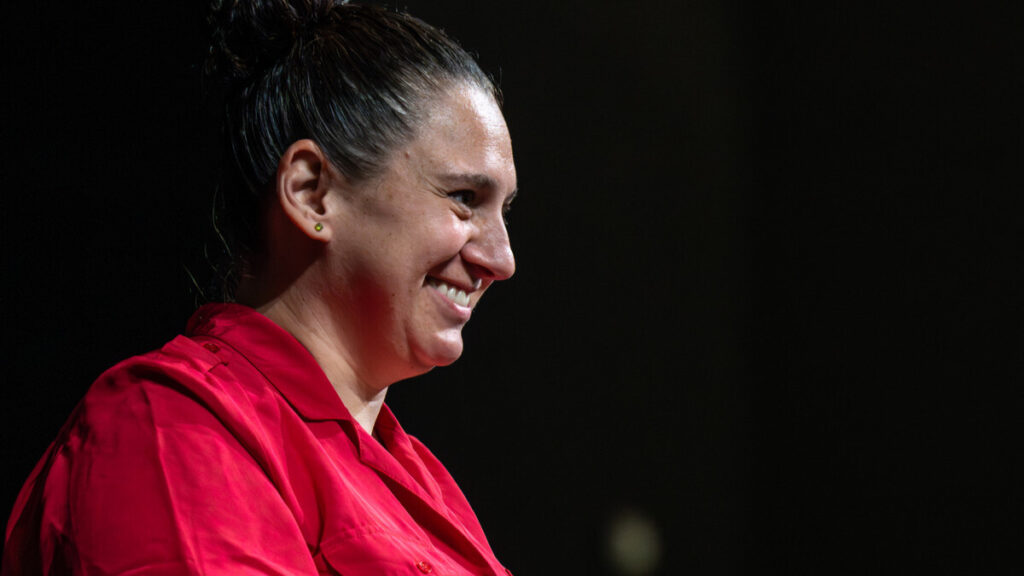 A close-up photo of a person wearing a bright red shirt and smiling to the right. They have dark hair pulled behind their head. The background is dark.