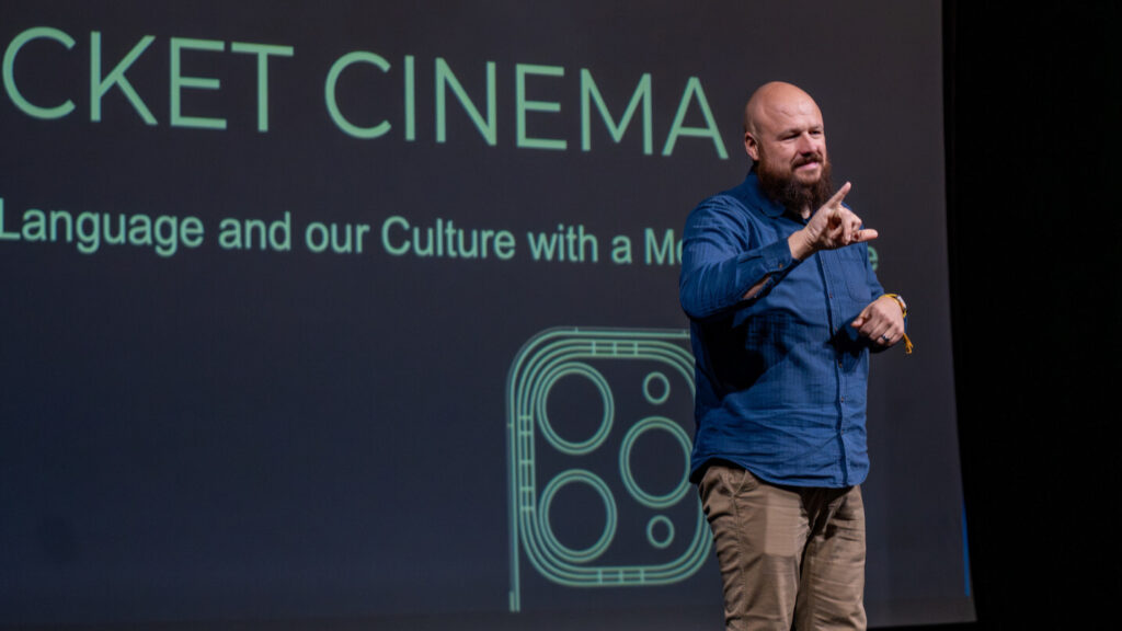 A photo of a person signing onstage to an unseen audience to the right. They are bald, with a dark brown beard. They wear a blue button-up shirt and slacked. Behind them is a projected presentation. Obscured text says "Pocket Cinema / [obscured] Language and our Culture with a Mobile Phone".