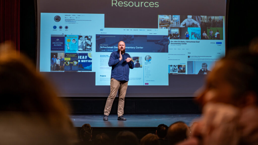 A photo from behind a crowd of a person signing onstage. They are bald, with a dark brown beard. They wear a blue button-up shirt and slacked. Behind them is a projected presentation. Text at the top says "Resources" and the screen has various screenshots of web resources.