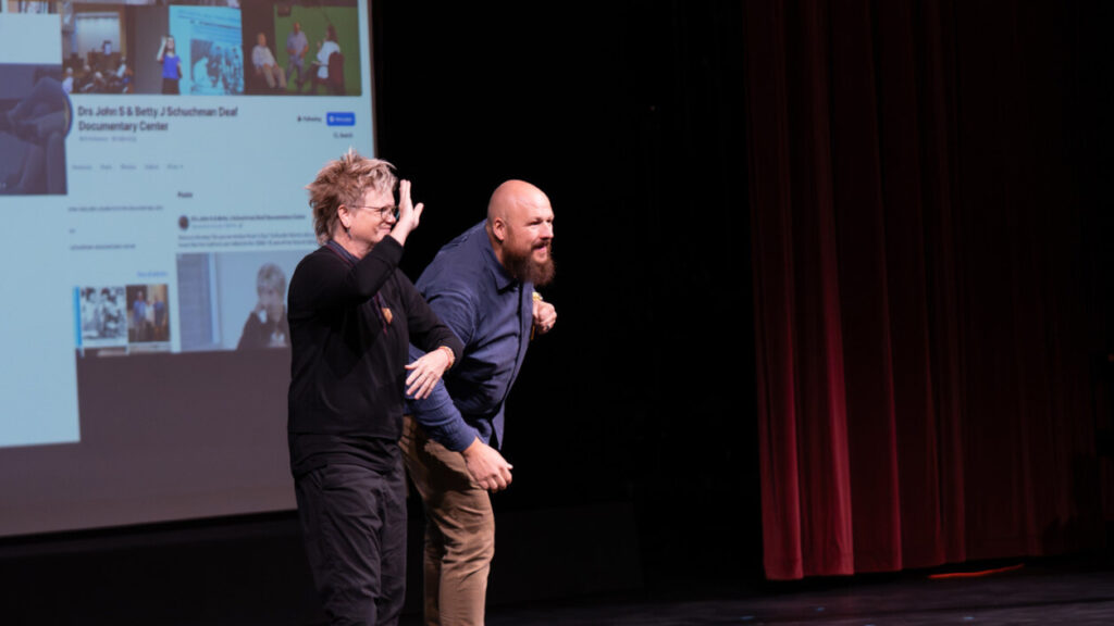 A photo of two people onstage looking to the right at an offscreen audience. They have a presentation behind them to the left. The person on the left appears to be an interpreter. The person on the right is leaning forward playfully.