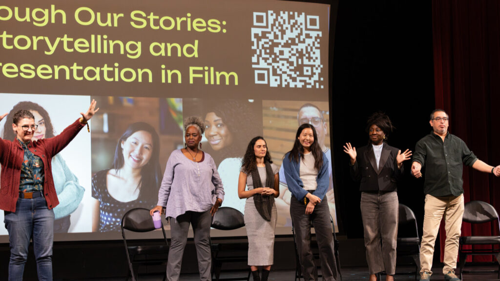 A photo of six standing people onstage addressing an unseen audience to the right. A projected screen behind has obscured text: "Through Our Stories: Storytelling and Representation in Film", as well as photos of the five people on the right. The person on the left is handwaving the audience.