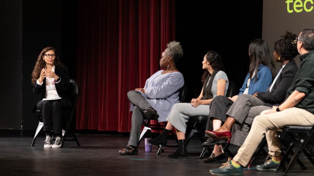 A photo of six panelists seated onstage. The five on the right are looking at the moderator on the left, who is signing a question.