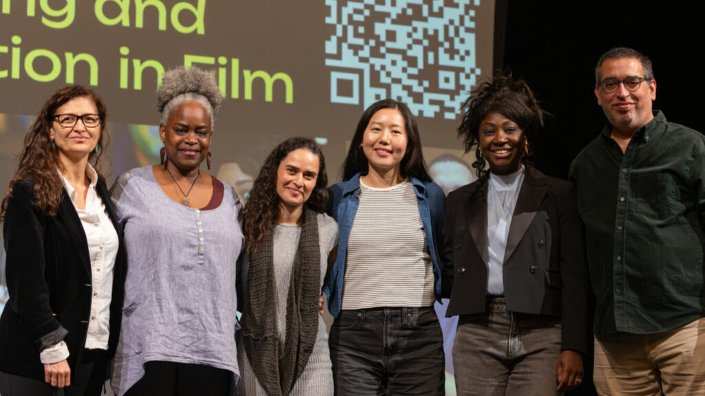 A photo of six people on a stage posing for the camera. They represent various identities and have different attire. A projected screen in the background has obscured text and a QR code.