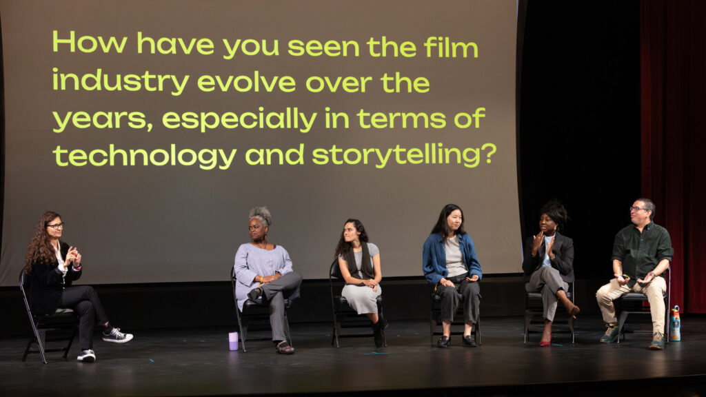 A photo of six panelists seated onstage. The moderator on the left and the panelist fifth from left are signing. In the back is a projected screen with text: How have you seen the film industry evolve over the years, especially in terms of technology and storytelling?".