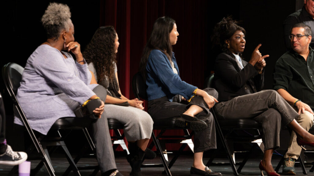 A photo of five seated panelists onstage. The panelist fourth from left is signing to an unseen audience to the right. The other panelists look on.
