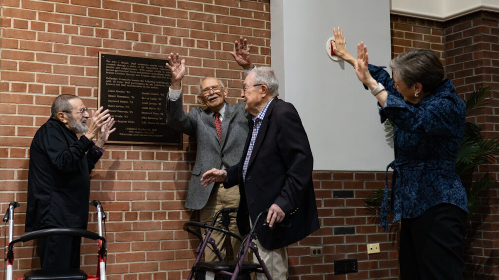 A photo of three seniors cheering next to a plaque on a brick wall. One (second from left) is facing the camera, while the others face each other. Another cheering person is visible on the right.