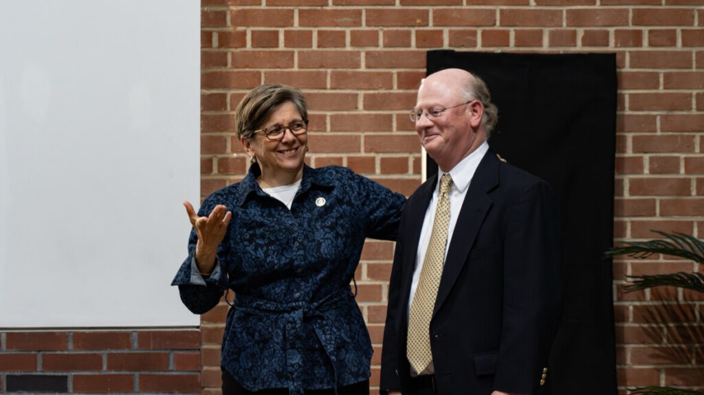 Two smiling people stand in front of an unseen crowd. The person on the left in a smart dress jacket has their arm around the other, who wears a suit and tie. A brick wall with presentation screens is visible in the background.